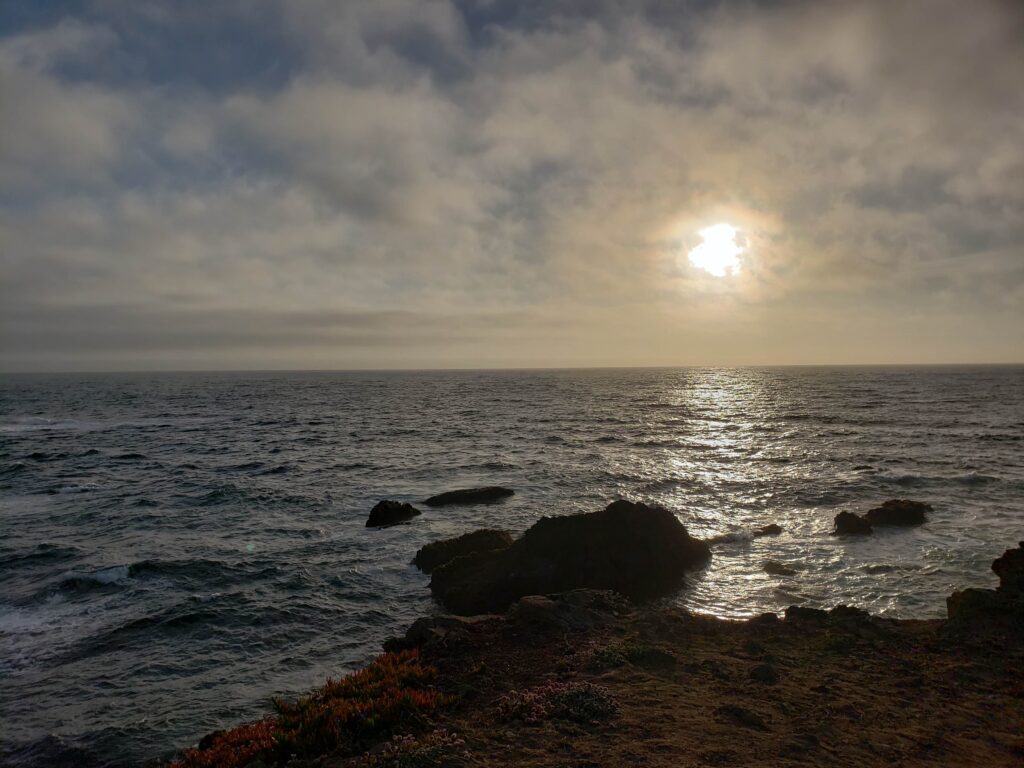 The sun sets through a foggy and cloud-filled horizon over the Pacific ocean which stops at the rocky coastline at the bottom of the picture.
