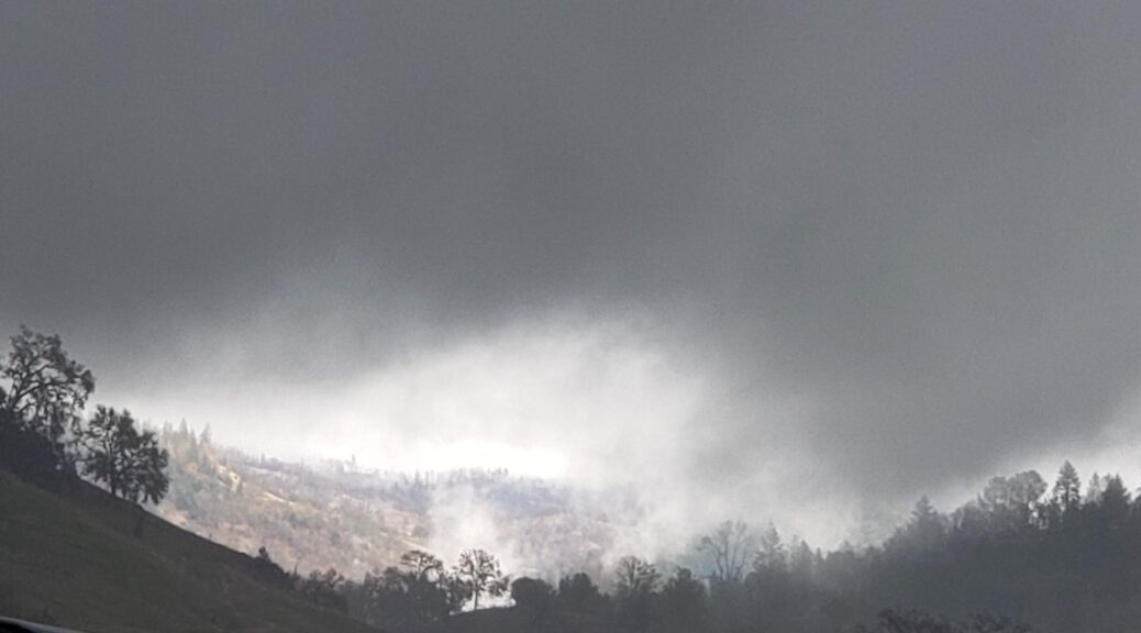 A landscape, dominated by dark gray clouds that fill the majority of this picture, though a small area is illuminated beyond the gloom, revealing a landscape of color and light beyond the darkness, and the tree line kept in silhouette by the gloom dominating the landscape.