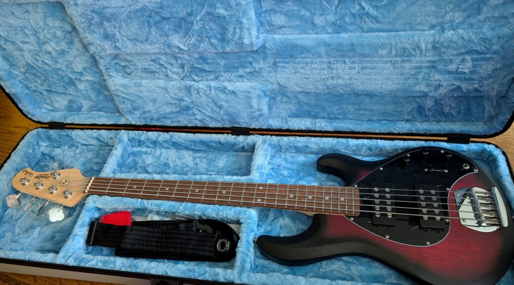 Sterling Stingray 5 HH with a sunburst finish in a light blue case.